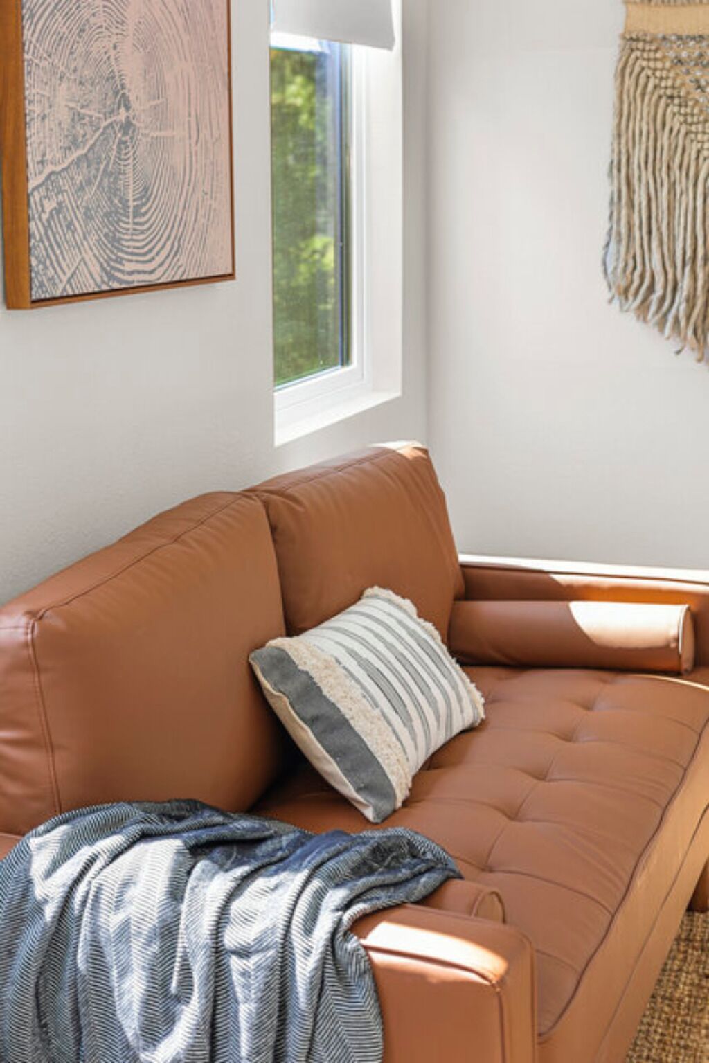 A tan leather sofa with a striped pillow and a blanket, a window with a view of greenery, and a wall-mounted tapestry.