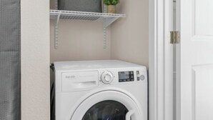 A compact laundry room with a front-loading washing machine, a shelf above it, and a door to the right. - Tiny Home  - (Cedar) (Dunsmuir)