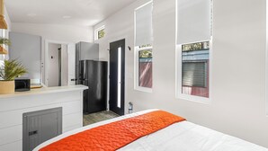 A modern bedroom with a bed, a built-in wardrobe, a kitchen area with a refrigerator, and a window with blinds. - Tiny Home   -  (Koa) (Dunsmuir)
