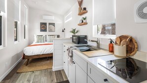 A compact, modern bedroom with a bed, bedside tables, a built-in microwave, and a small kitchenette. - Tiny Home   -  (Koa) (Dunsmuir)