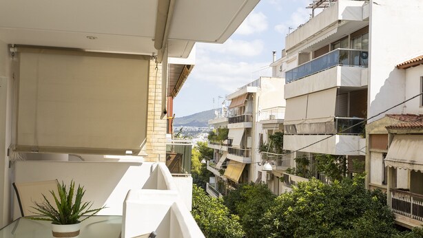 Un balcon agrémenté d’un pot de fleurs, donnant sur un quartier résidentiel composé de plusieurs immeubles et offrant une vue lointaine sur une montagne.