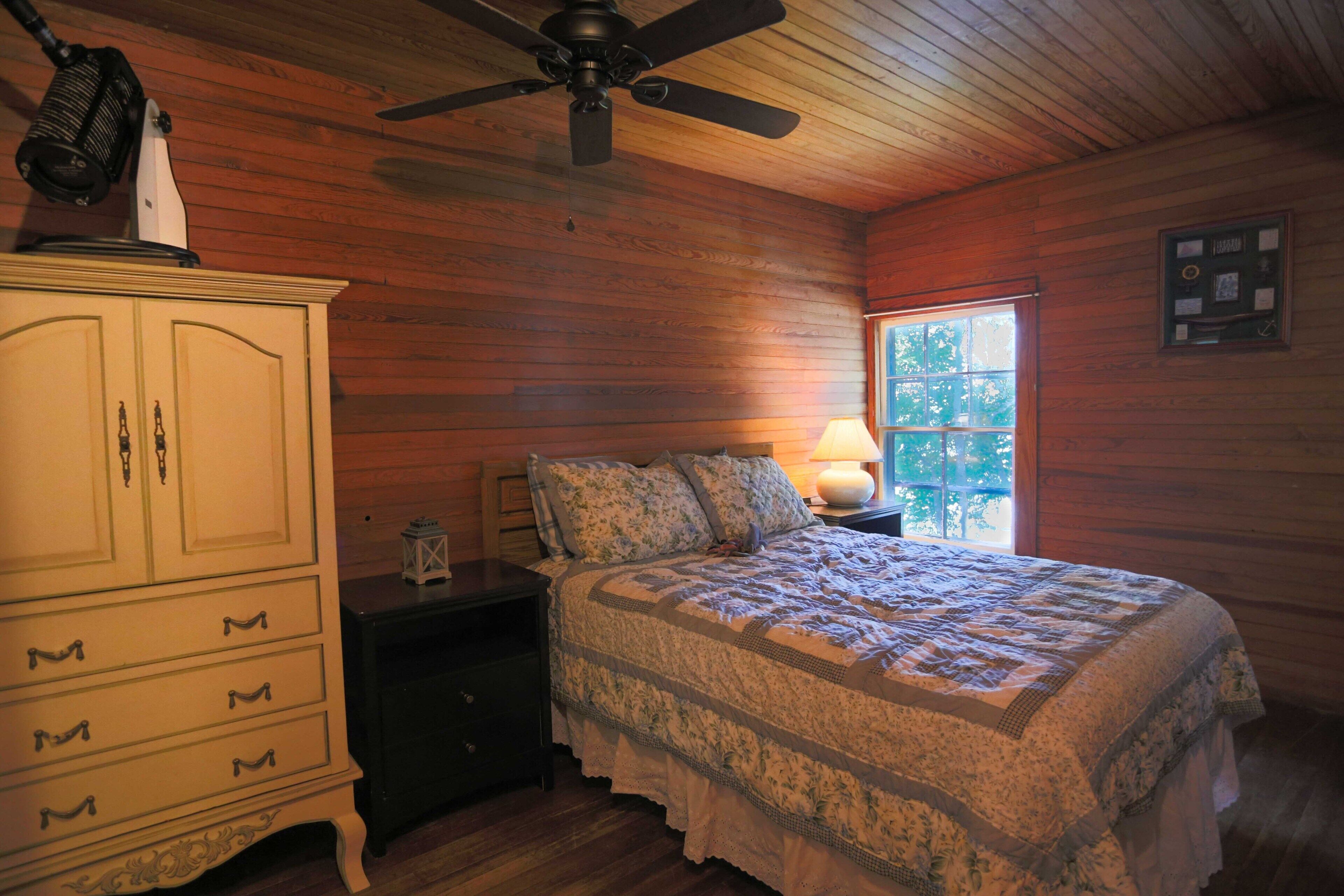 Ein Schlafzimmer mit Holzboden, einem Bett mit floralem Überwurf, einem Nachttisch mit Lampe und einer Kommode mit Schubladen.