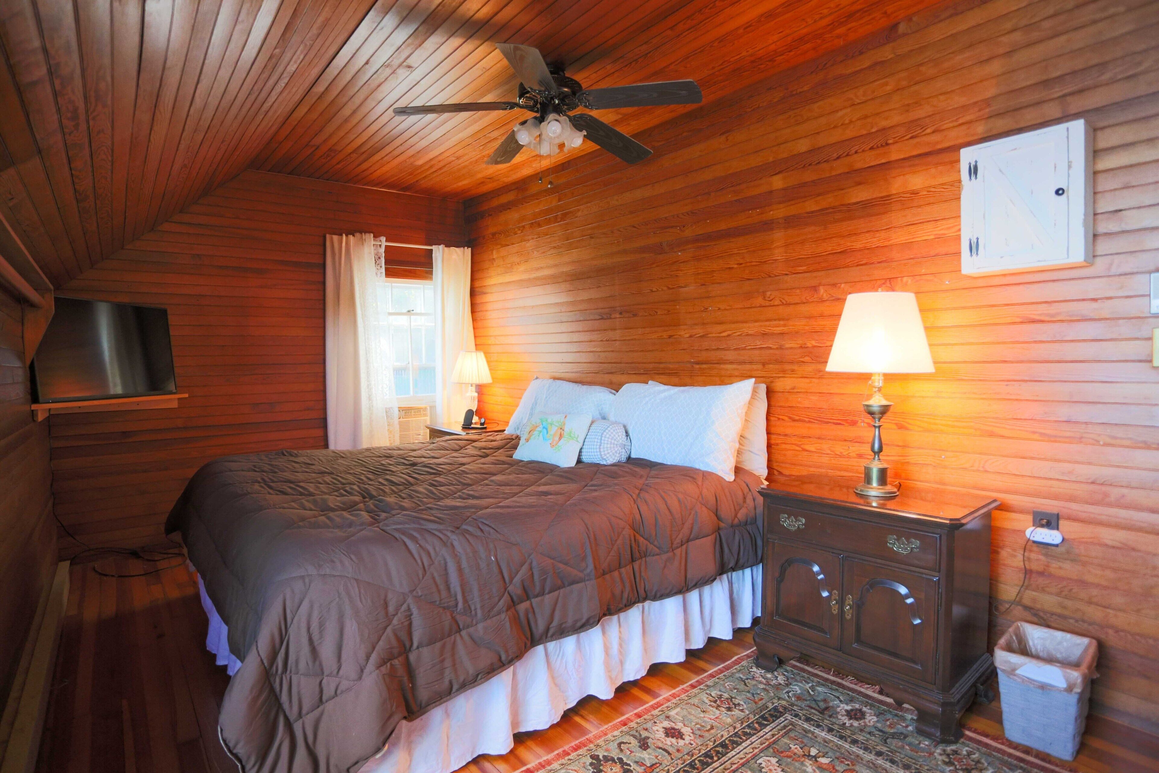 Un dormitorio acogedor con techo de madera, una cama con colcha marrón, dos lámparas de noche, un televisor en un estante y un pequeño cubo de basura.