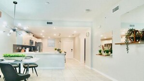 A modern kitchen with a central island, dining area with chairs, and a hallway with a plant shelf.