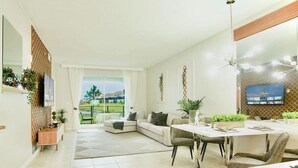 A modern living room with a sofa, dining table, and TV. There is a balcony with plants and a view of the outdoors.