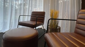 Une pièce dotée d’une grande fenêtre, d’un lustre, d’un canapé en cuir marron, d’un fauteuil en cuir marron et d’une petite table sur laquelle est posé un vase rempli de plantes.