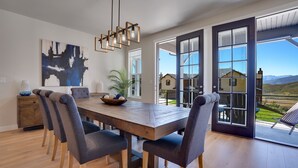 A dining area with a wooden table, grey upholstered chairs, a hanging light fixture, and a large window with a view of a house and mountains.