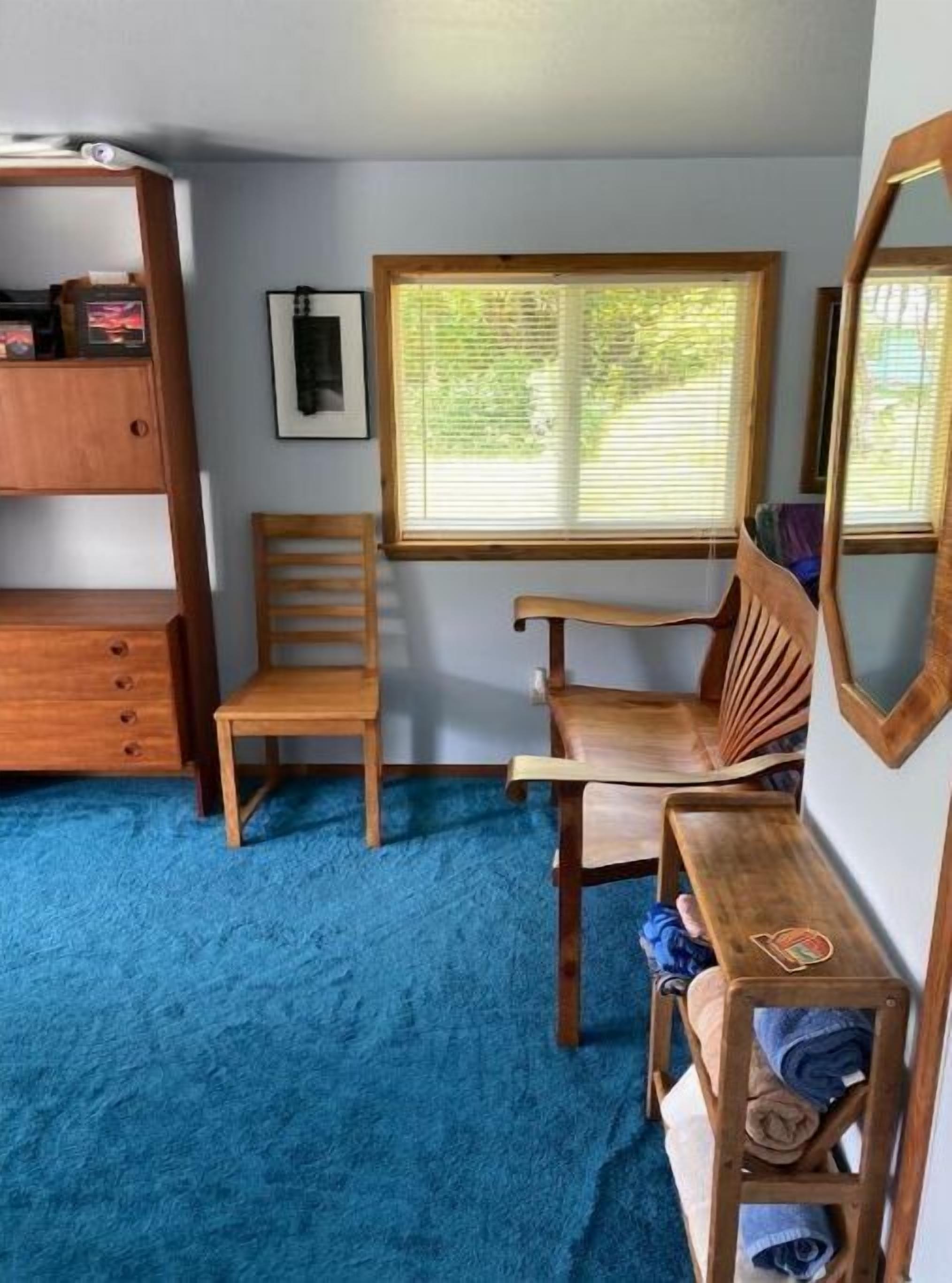 A room with a blue carpet, a wooden chair, a wooden desk, a mirror, and a window with blinds.