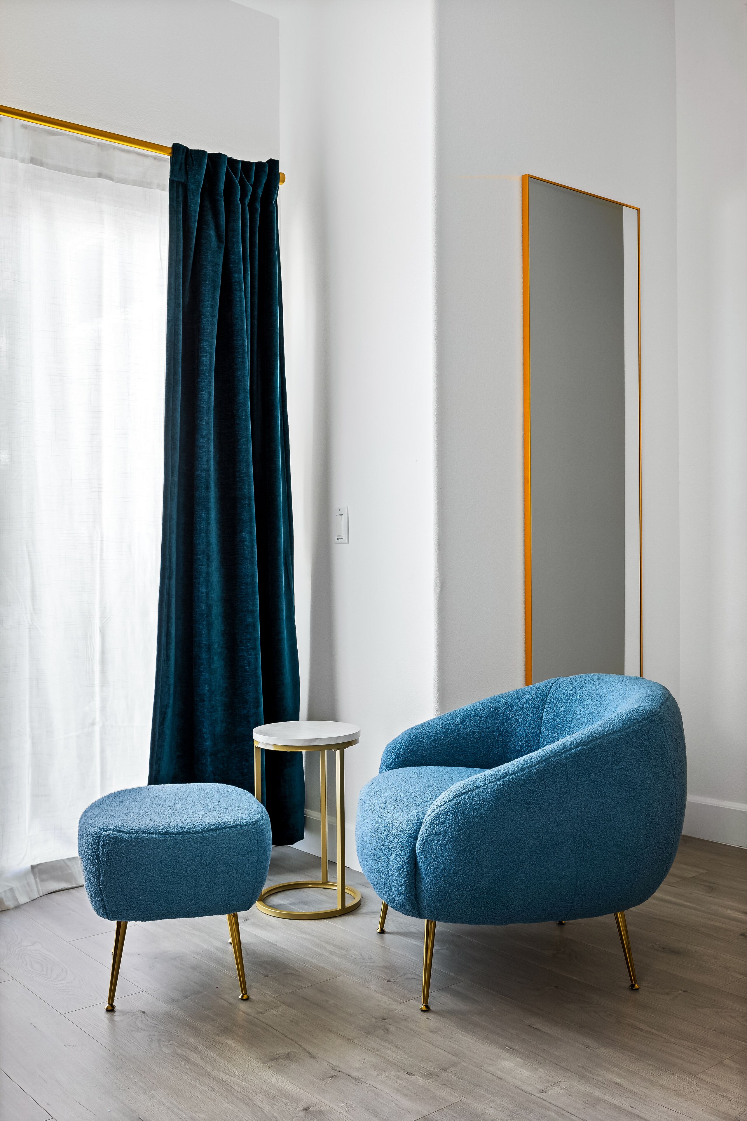 Ein modernes Zimmer mit blauem Ohrensessel, kleinem runden Tisch und großem Spiegel.