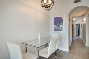 Een moderne eetruimte met een glazen tafel en witte stoelen, een kroonluchter en een ingelijst kunstwerk aan de muur.