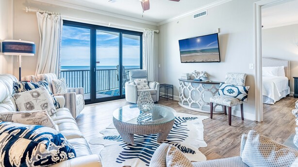 A spacious living room with a large glass coffee table, a sofa set, a dining area, and a view of the ocean through a sliding glass door.