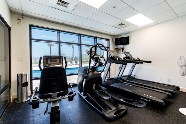 Une salle de sport équipée de tapis de course, d’un vélo elliptique et d’un vélo d’appartement, de grandes fenêtres offrant une vue sur les palmiers et d’un téléviseur fixé au mur.