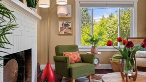 A cozy living room with a green armchair, a fireplace, a large window with a view of greenery, and a vase with red flowers. - Charming 1920s Olympia home with game room, fenced yard, blocks from Capitol (Olympia)