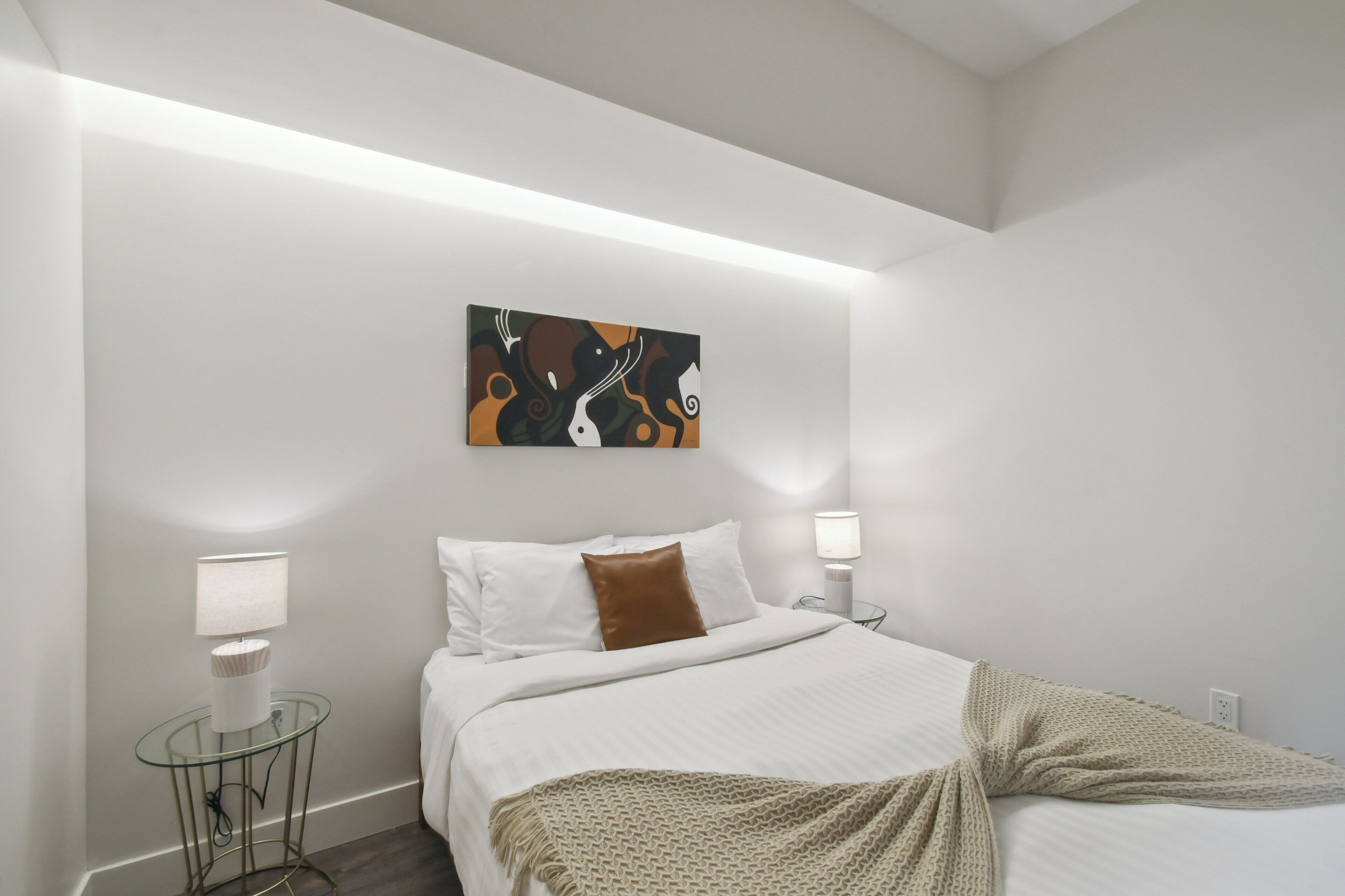 A modern bedroom with a bed, white walls, a painting, two bedside lamps, and a glass table.