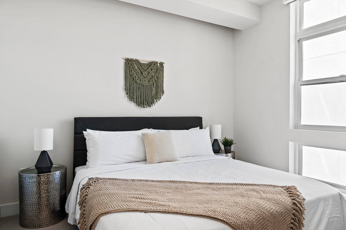 Une chambre à coucher avec un lit, du linge de lit blanc, une tête de lit sombre, une décoration murale, une lampe de chevet et une petite plante.