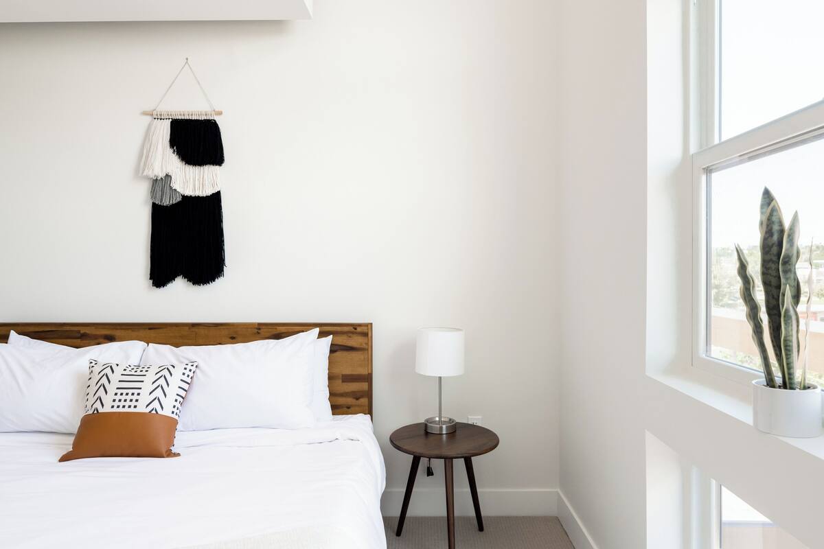 Une chambre d’hôtel avec une tête de lit en bois, du linge de lit blanc, une lampe de chevet, une petite table ronde et une vue sur l’extérieur par une fenêtre.