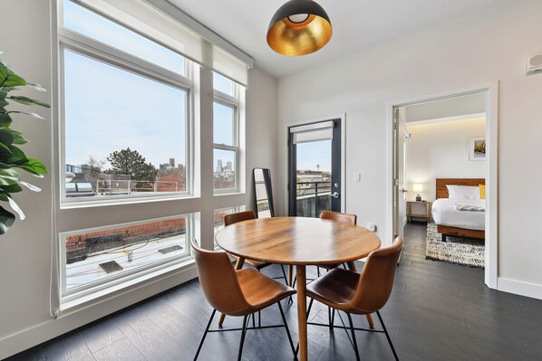 Une chambre moderne avec une table ronde en bois, des fauteuils en cuir, de grandes fenêtres, un lit et une vue sur la ville.