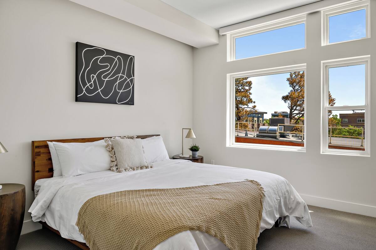 Une chambre avec un grand lit, une fenêtre donnant sur des immeubles et une œuvre d’art murale.