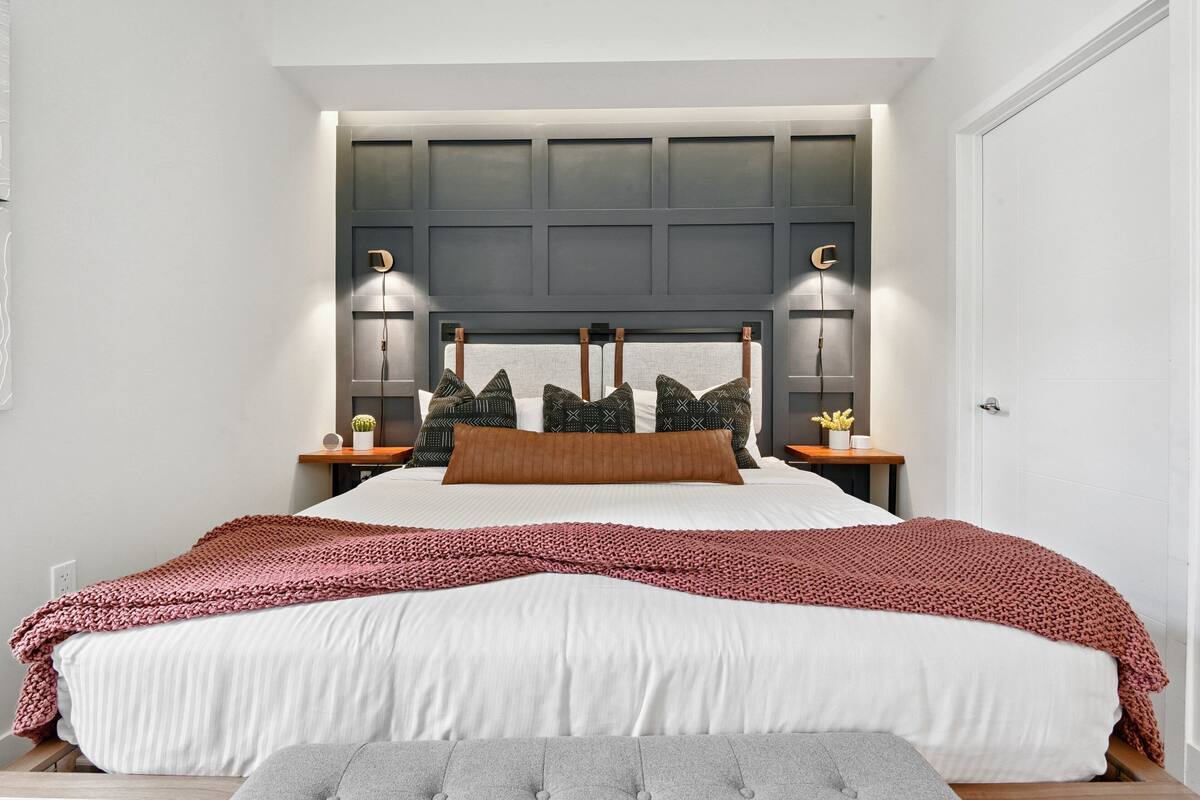 Une chambre à coucher avec un grand lit, une tête de lit dotée d’un éclairage intégré, des tables de chevet avec des lampes et un banc gris.