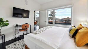 A modern hotel room with a large bed, a TV mounted on the wall, a desk with a chair, and a window with a view of buildings. - Espadin Condos II (Denver)