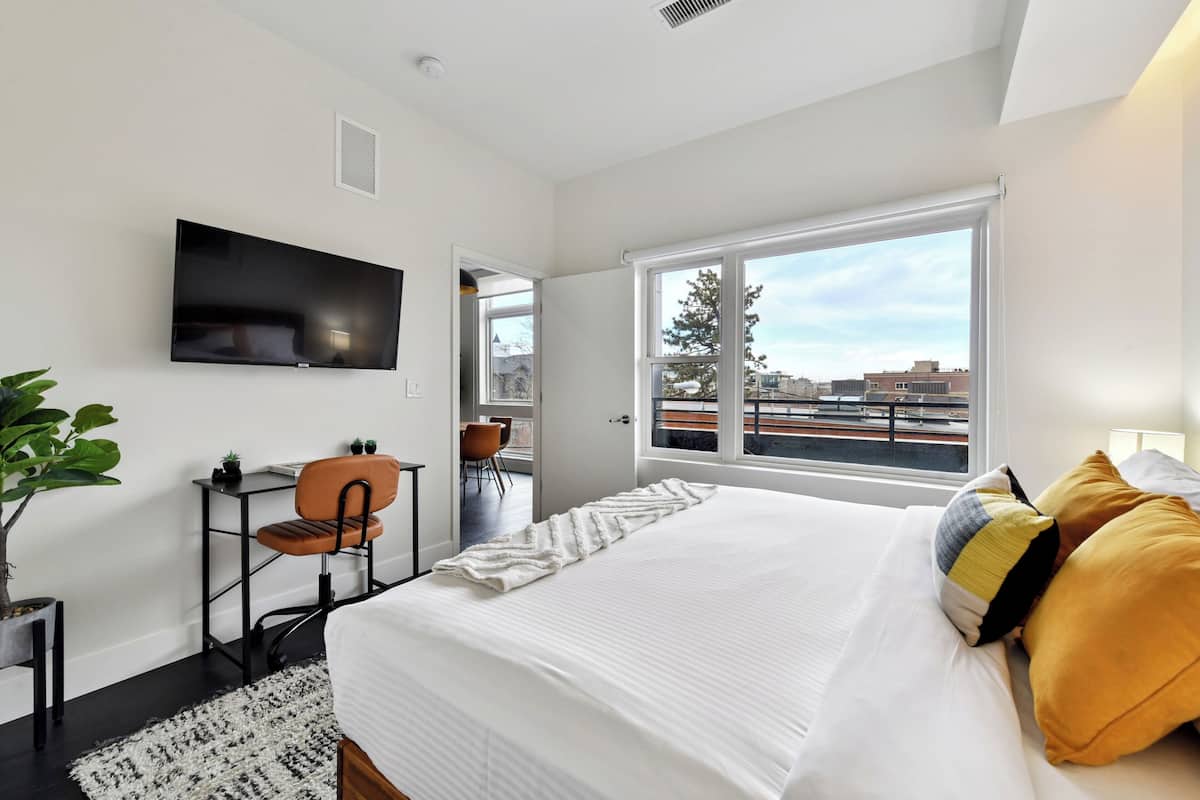 Une chambre d’hôtel moderne dotée d’un grand lit, d’un téléviseur fixé au mur, d’un bureau avec une chaise et d’une fenêtre donnant sur des immeubles.