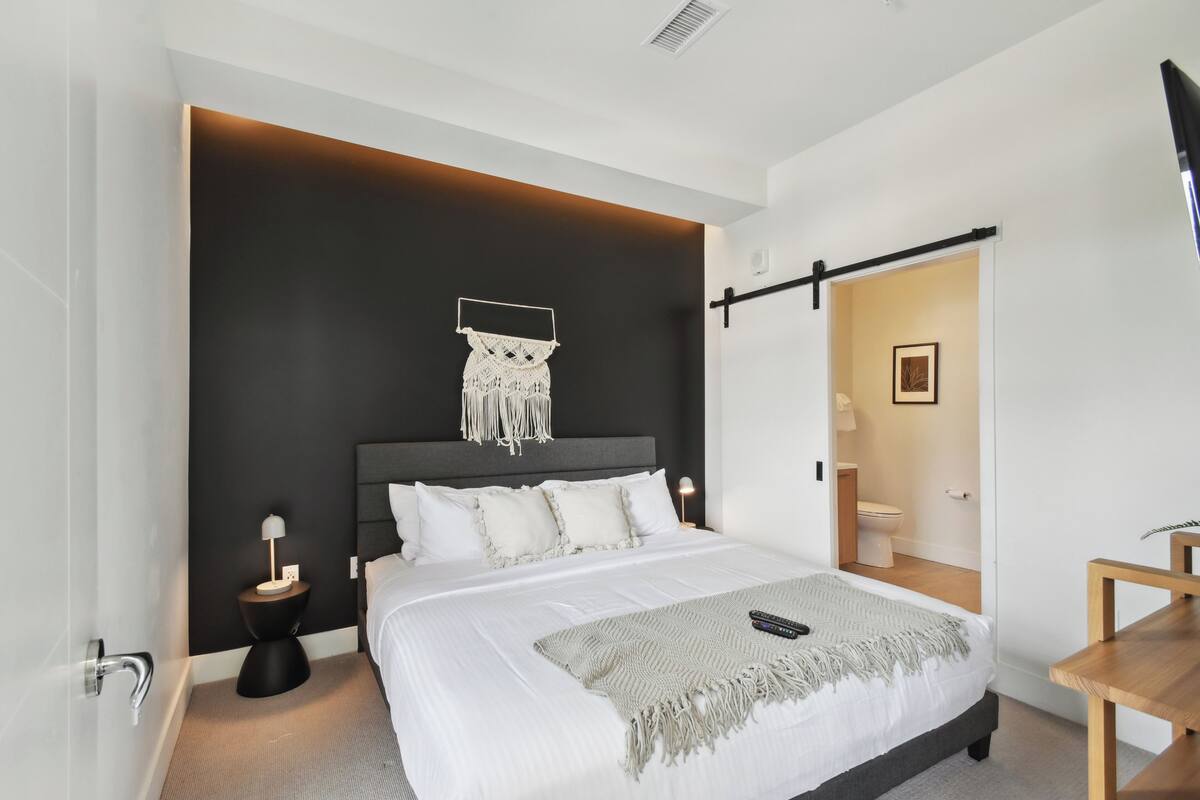 Une chambre à coucher moderne avec un grand lit, un mur d’accent de couleur sombre, une décoration murale en macramé, une table de chevet avec une lampe et une chaise en bois.