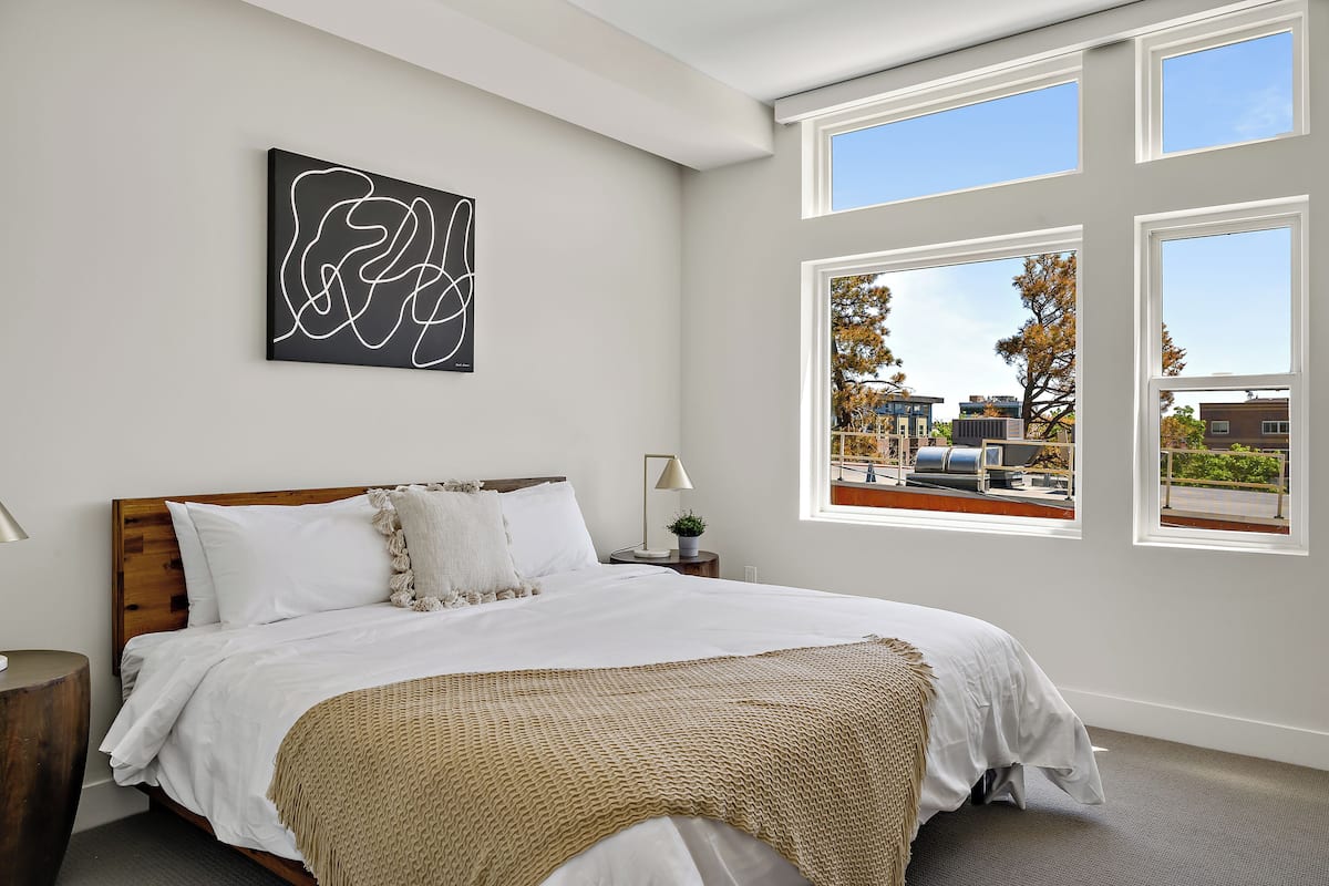 Une chambre avec un grand lit, une fenêtre donnant sur des immeubles et une œuvre d’art abstraite accrochée au mur.