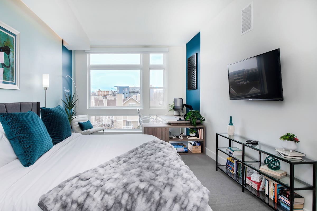 Ein modernes Schlafzimmer mit einem großen Bett, Blick auf die Stadt, einem Fernseher und einem Bücherregal.