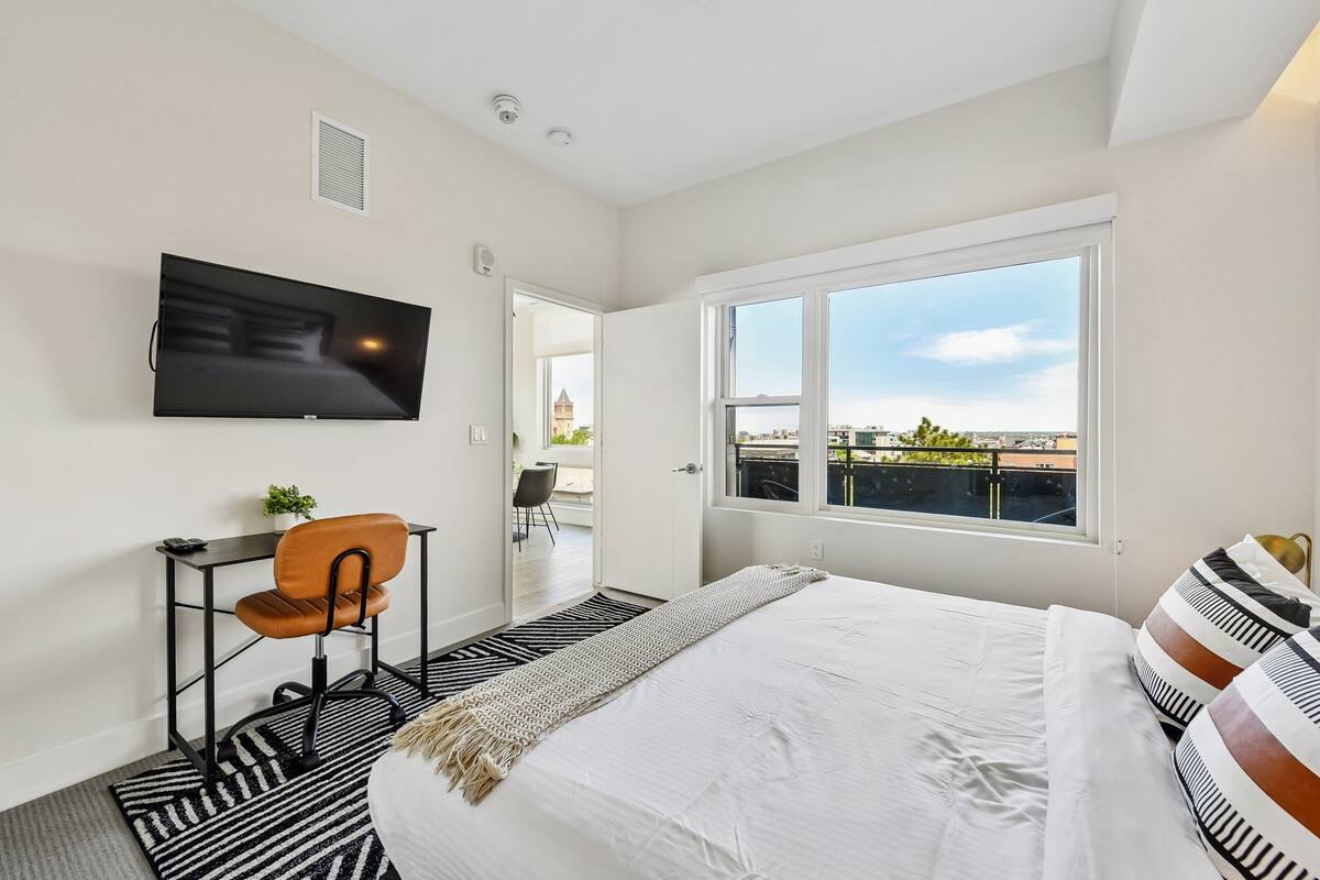 Une chambre à coucher comprenant un lit, un téléviseur, un bureau avec une chaise, une fenêtre donnant sur un paysage agréable et une porte menant à un balcon.