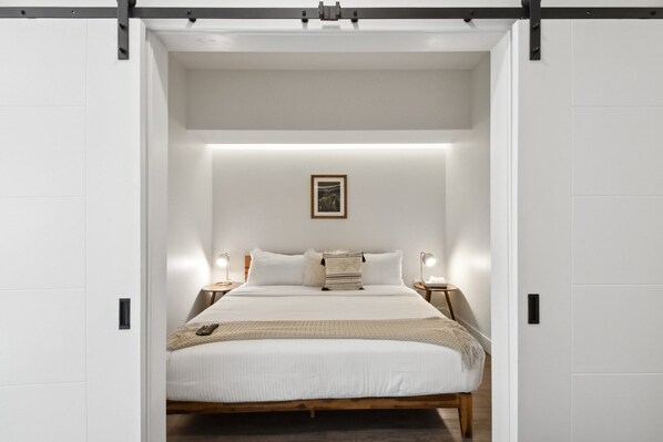 A minimalist bedroom with a bed centered in the room, flanked by bedside tables with lamps. - Espadin Condos I (Denver)