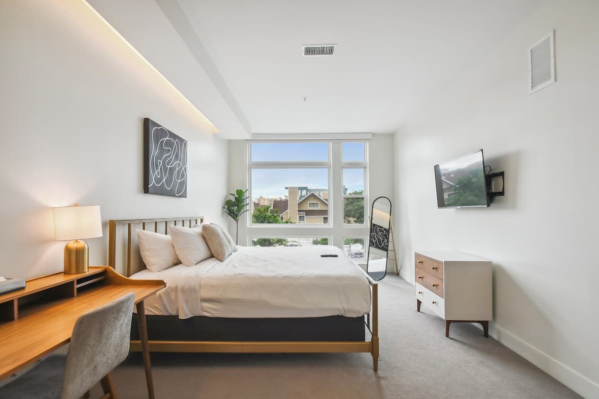 Ein modernes Schlafzimmer mit Bett, Schreibtisch, Stuhl, Kommode und einem großen Fenster mit Blick auf die Stadt.