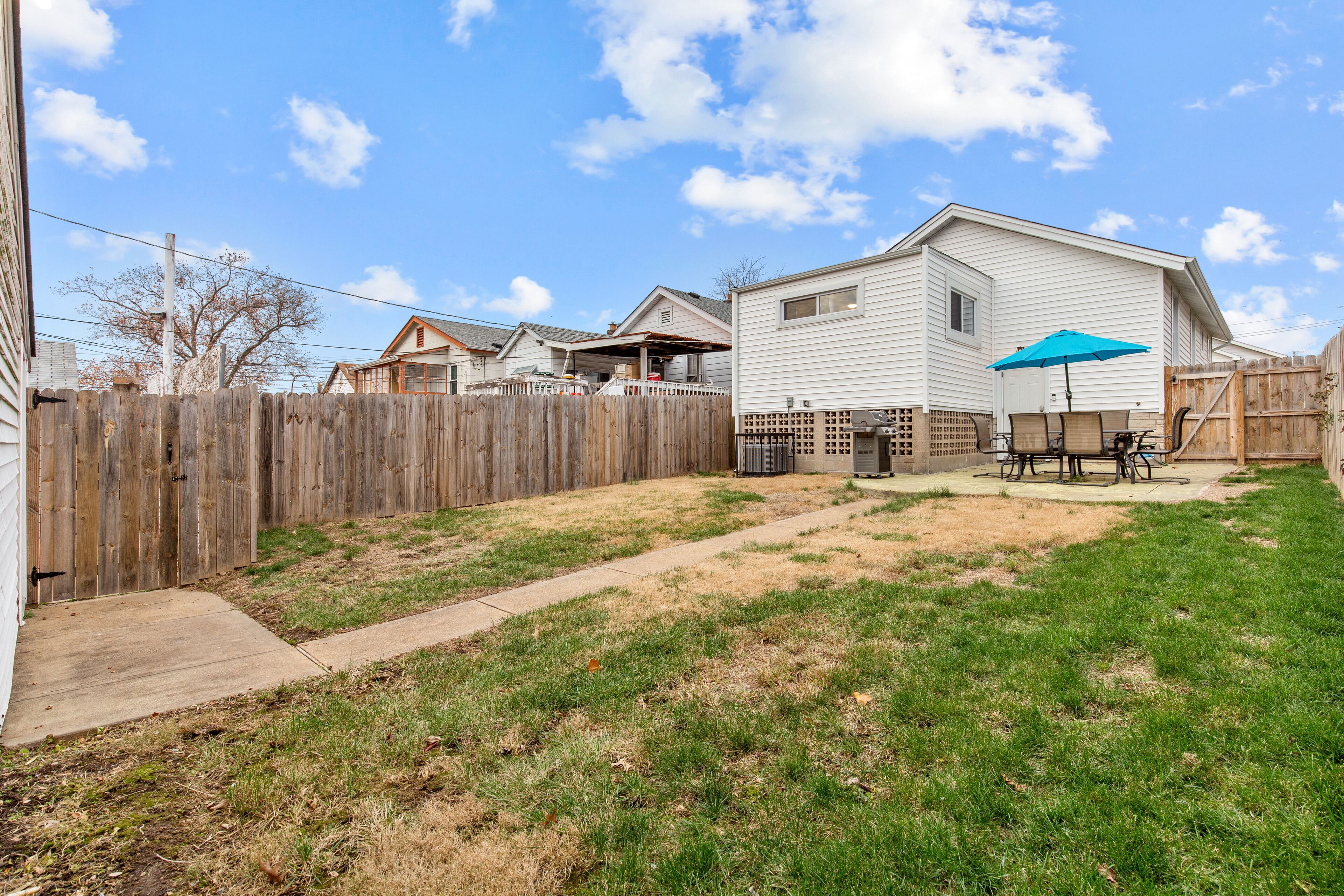 A residential backyard with a wooden fence, a concrete pathway, and a patio area with a table and chairs.