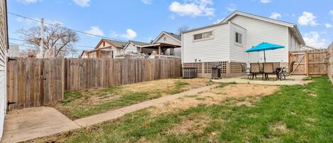 A residential backyard with a wooden fence, a concrete pathway, and a patio area with a table and chairs.