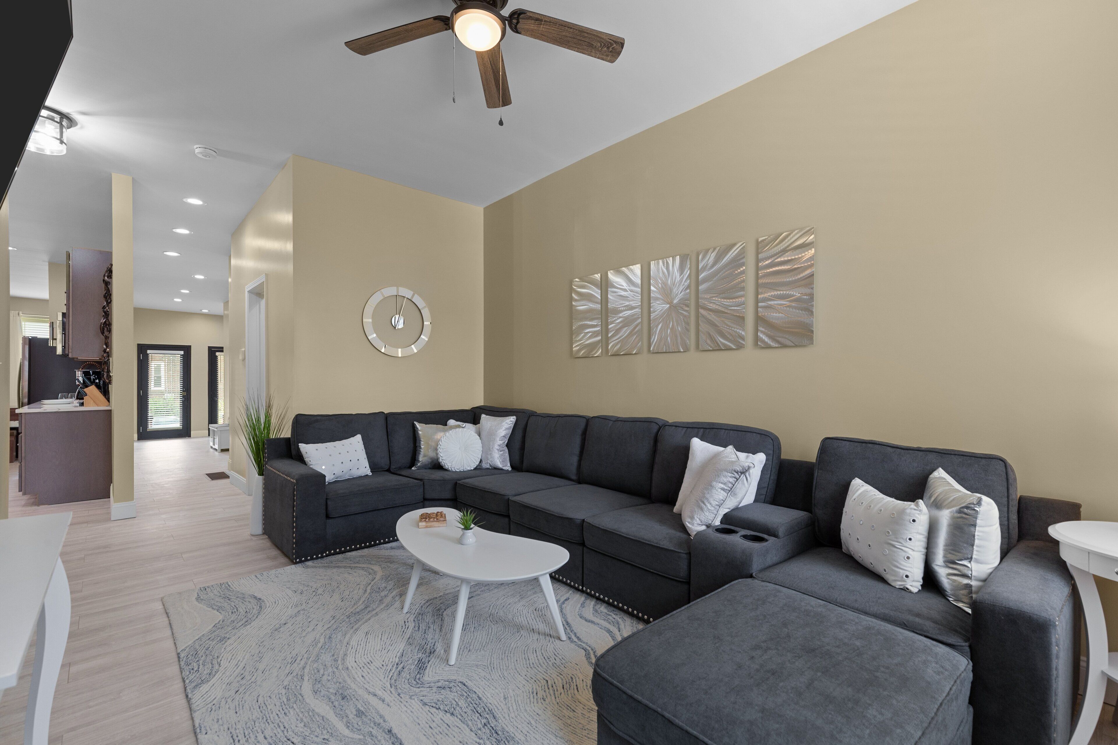 Ein modernes Wohnzimmer mit Ecksofa, Couchtisch und Deckenventilator. Im Hintergrund ist eine Küchenzeile zu sehen.