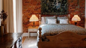 Ein Schlafzimmer mit einem hölzernen Bett, zwei Nachttischlampen, einem Nachttisch, einer Kommode, einem großen Gemälde an der Backsteinwand und einer Chandelier.