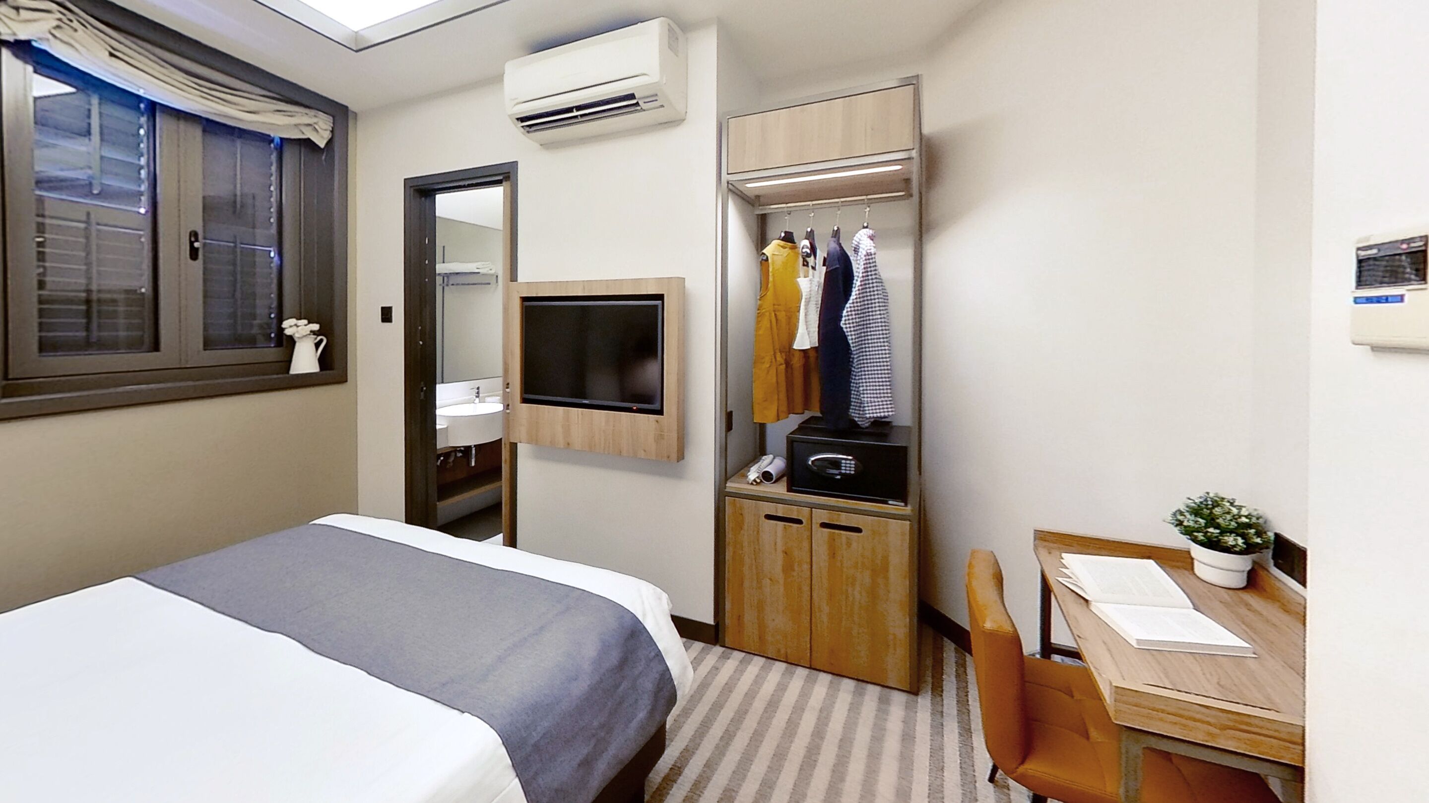 A hotel room with a bed, a desk with a chair, a mirror, a TV, a wardrobe, and an air conditioning unit.