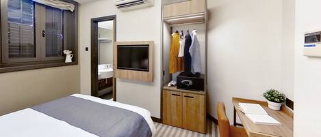 A hotel room with a bed, a desk with a chair, a mirror, a TV, a wardrobe, and an air conditioning unit.