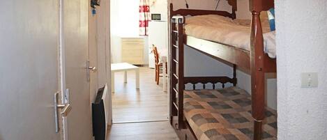 Un couloir étroit avec une porte en bois, un banc et un lit superposé doté d’un matelas à motifs.