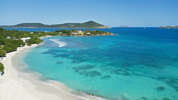 Eine Küstenlandschaft mit Strand, klarem türkisblauem Wasser und einer kleinen Insel mit Gebäuden in der Ferne.