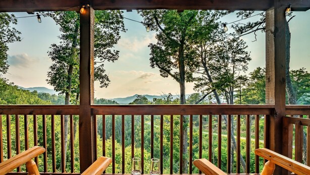 Una terraza de madera con vista a un bosque y montañas. Hay dos sillas y una mesa con vasos encima.