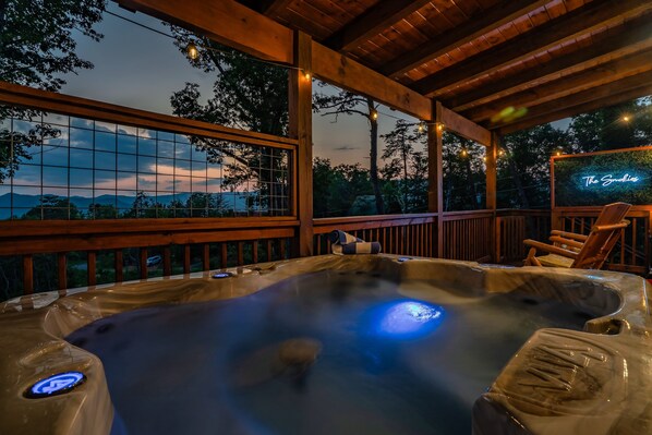 A hot tub with a view of the mountains, surrounded by wooden deck and chairs.