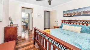 Une chambre à coucher comprenant un lit, un ventilateur de plafond, une tête de lit en bois, une table de chevet, une lampe et un tableau représentant un paysage urbain.