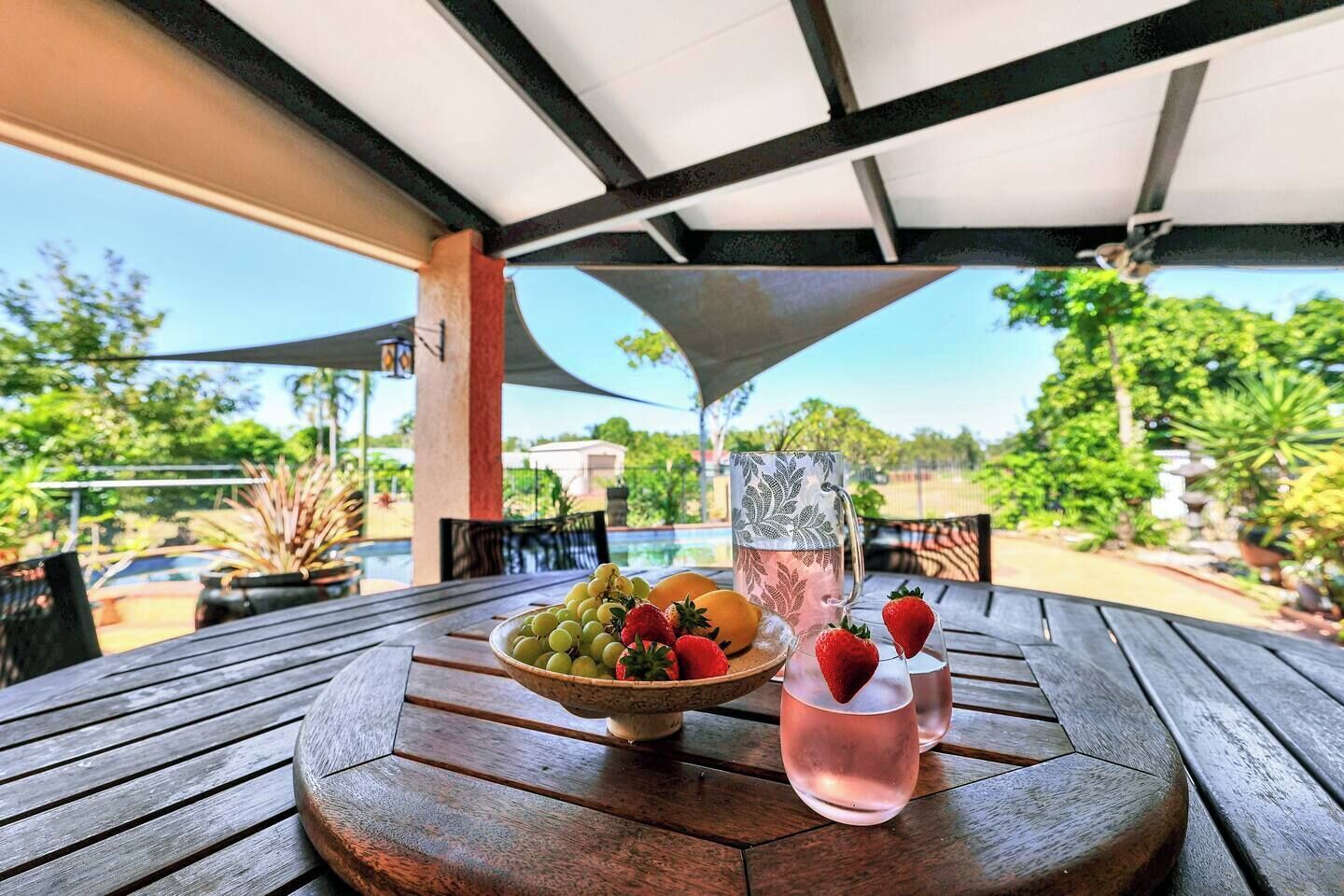 A wooden table with a bowl of fruit, a pitcher, and glasses on an outdoor patio with a pool view.