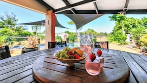 Une table en bois avec un saladier de fruits, une carafe et des verres, sur une terrasse extérieure donnant sur une piscine.