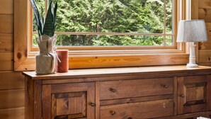 L’intérieur d’un chalet en bois, avec une grande fenêtre, une plante, une bougie et une lampe posée sur une commode en bois.
