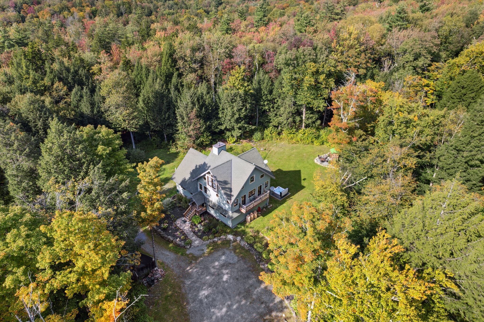 An aerial view of a large, two-story house surrounded by dense forest, with a visible driveway and a small pond.