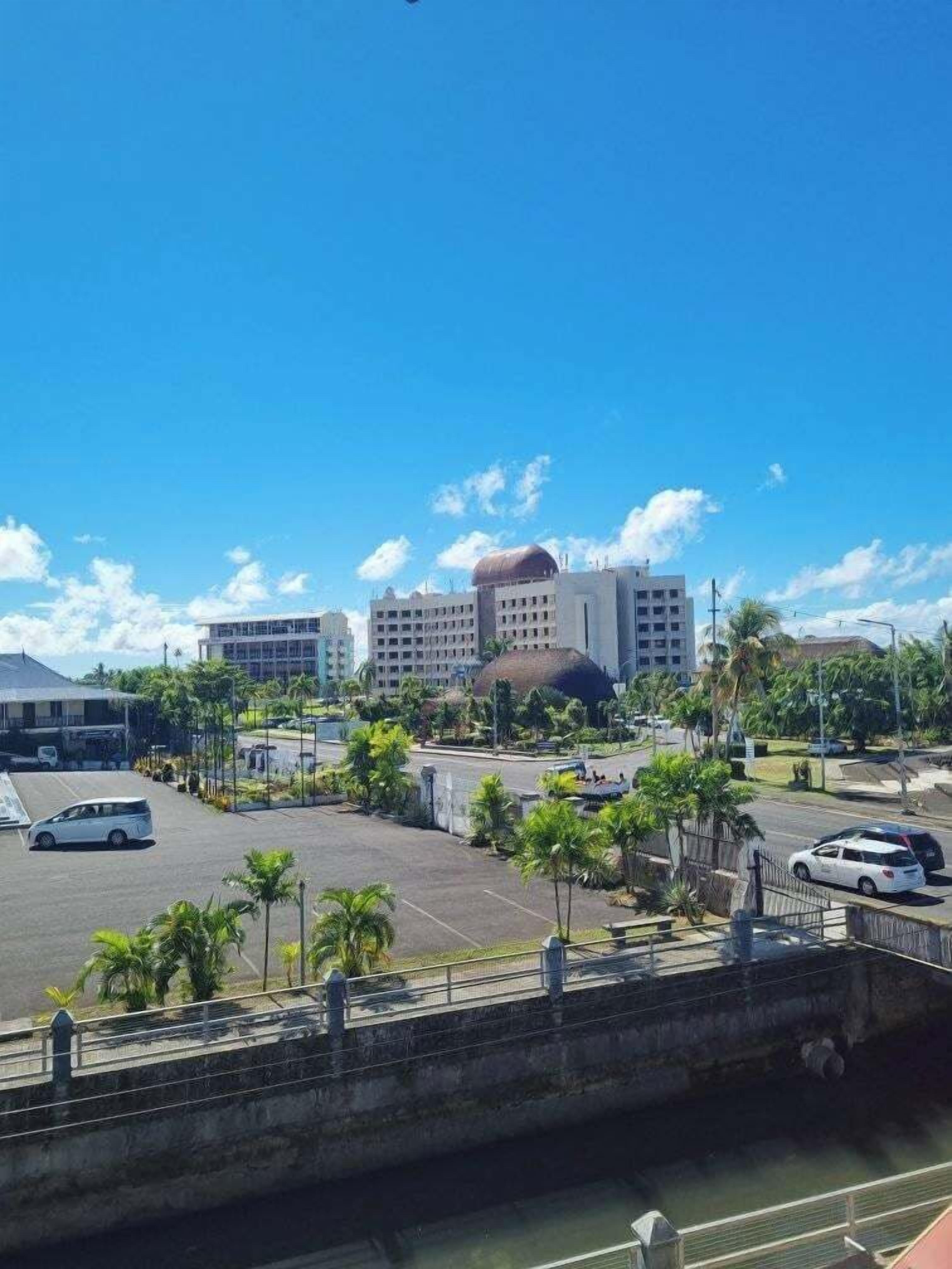 Un estacionamiento con varios coches, un puente y edificios al fondo.