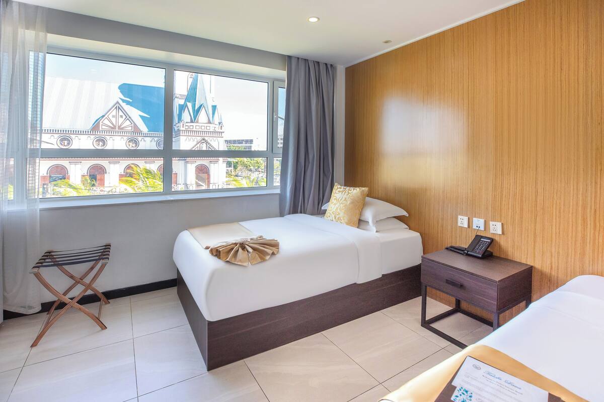 A hotel room with a bed, a nightstand, a small table, and a window with a view of a building.