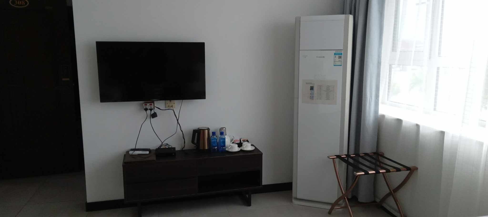 Una habitación con una televisión de pantalla plana en la pared, una mesita con una tetera y tazas, un refrigerador y una ventana con cortinas.