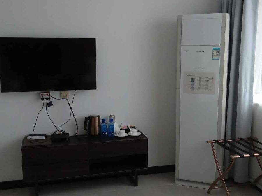 Una habitación con una televisión de pantalla plana en la pared, una mesita con una tetera y tazas, un refrigerador y una ventana con cortinas.